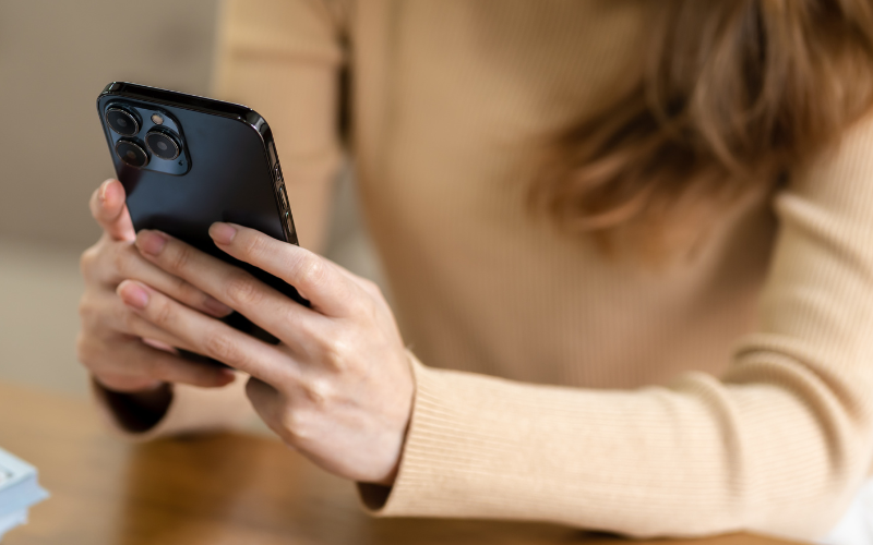 Person holding a black smartphone at a table, wearing a beige sweater.