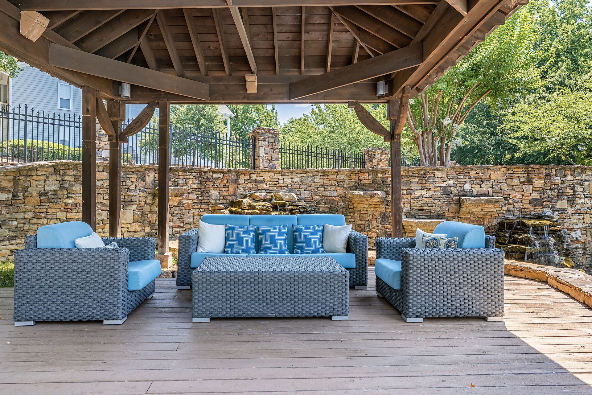 Outdoor seating area with blue cushions under a wooden gazebo at Marquis at Sugarloaf, offers apartments for rent near Sugarloaf Mills. Stone wall and trees in the background.