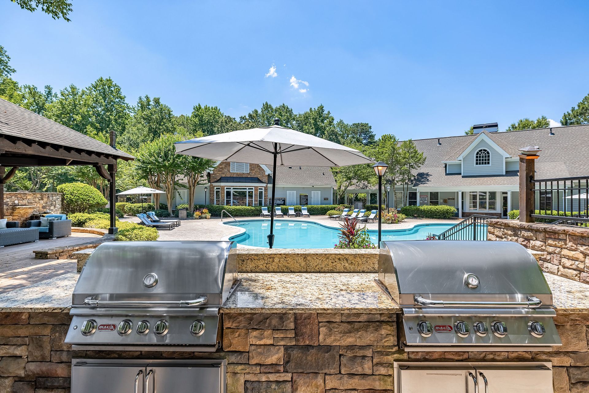 Two stainless steel grills on a brick countertop overlooking a swimming pool, patio, and building under a blue sky at Marquis at Sugarloaf, offers apartments for rent near Sugarloaf Mills.