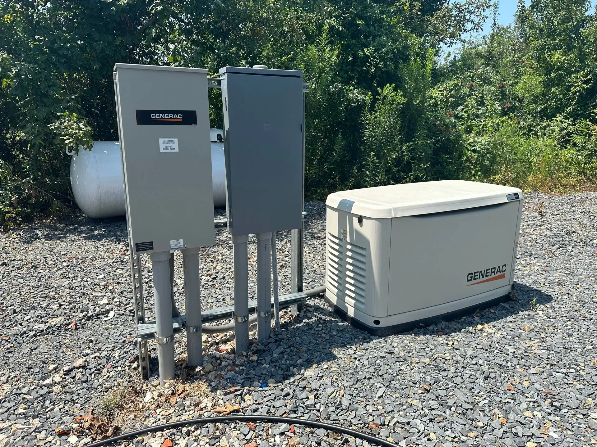 Outdoor electrical panel, generator, and propane tank on gravel. Green foliage in background under a blue sky.
