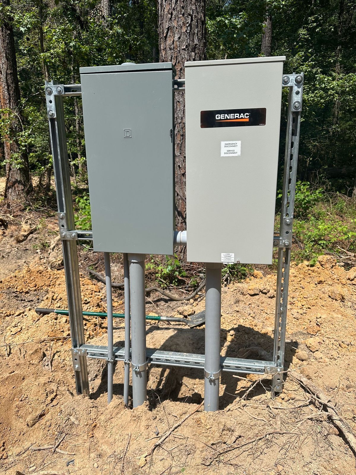 Two metal electrical boxes on poles, mounted on a metal frame, in a dirt and wooded setting.