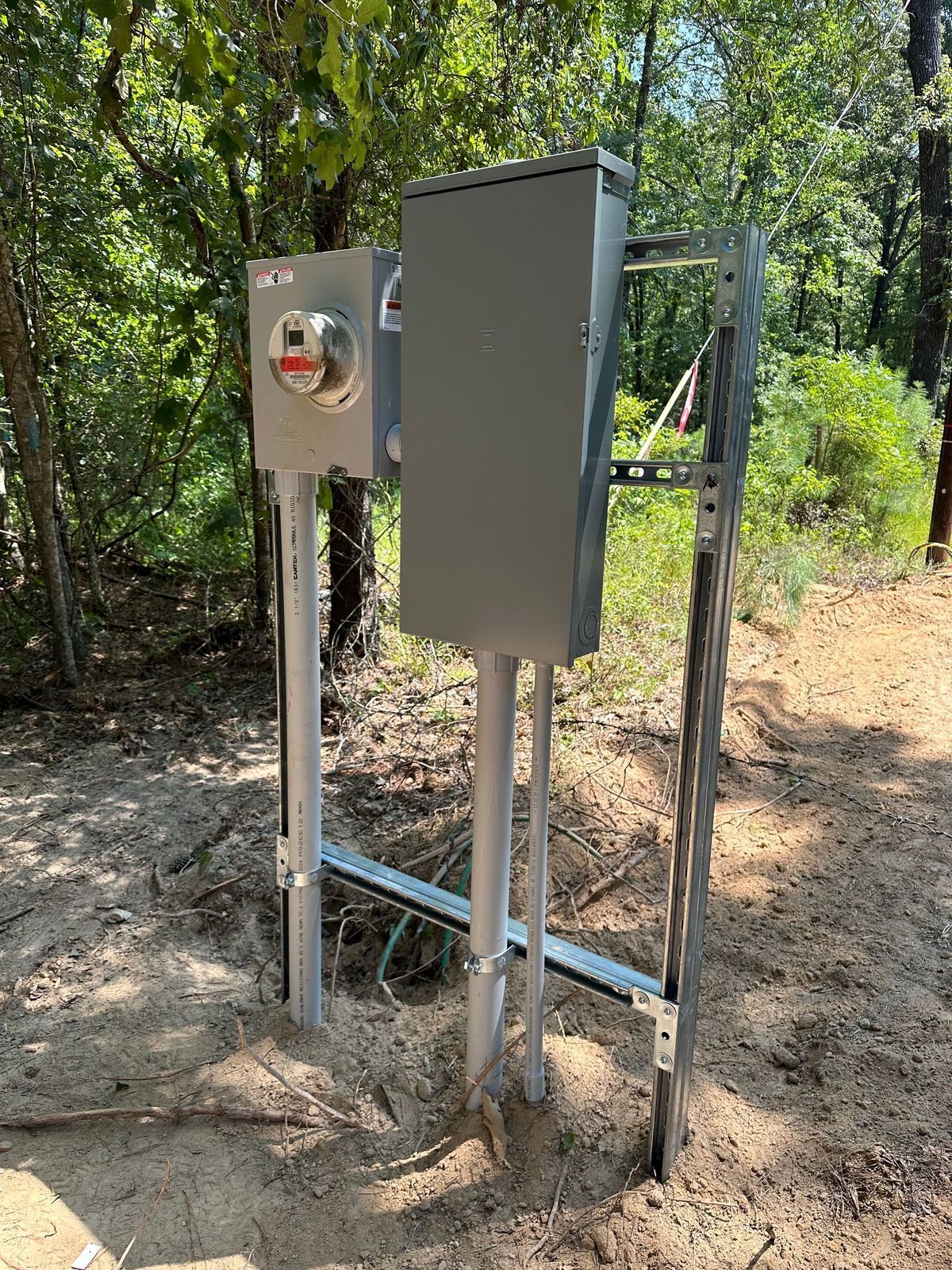 Outdoor electrical panel and meter, gray metal, installed on posts in a wooded area.