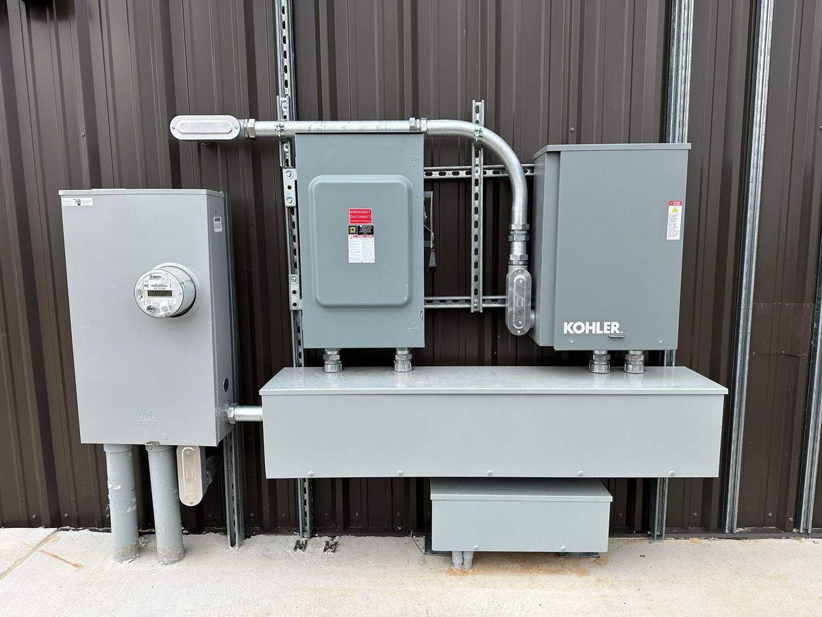 Gray electrical panel and boxes mounted on a dark wall. Includes a meter, circuit breaker, and generator panel.