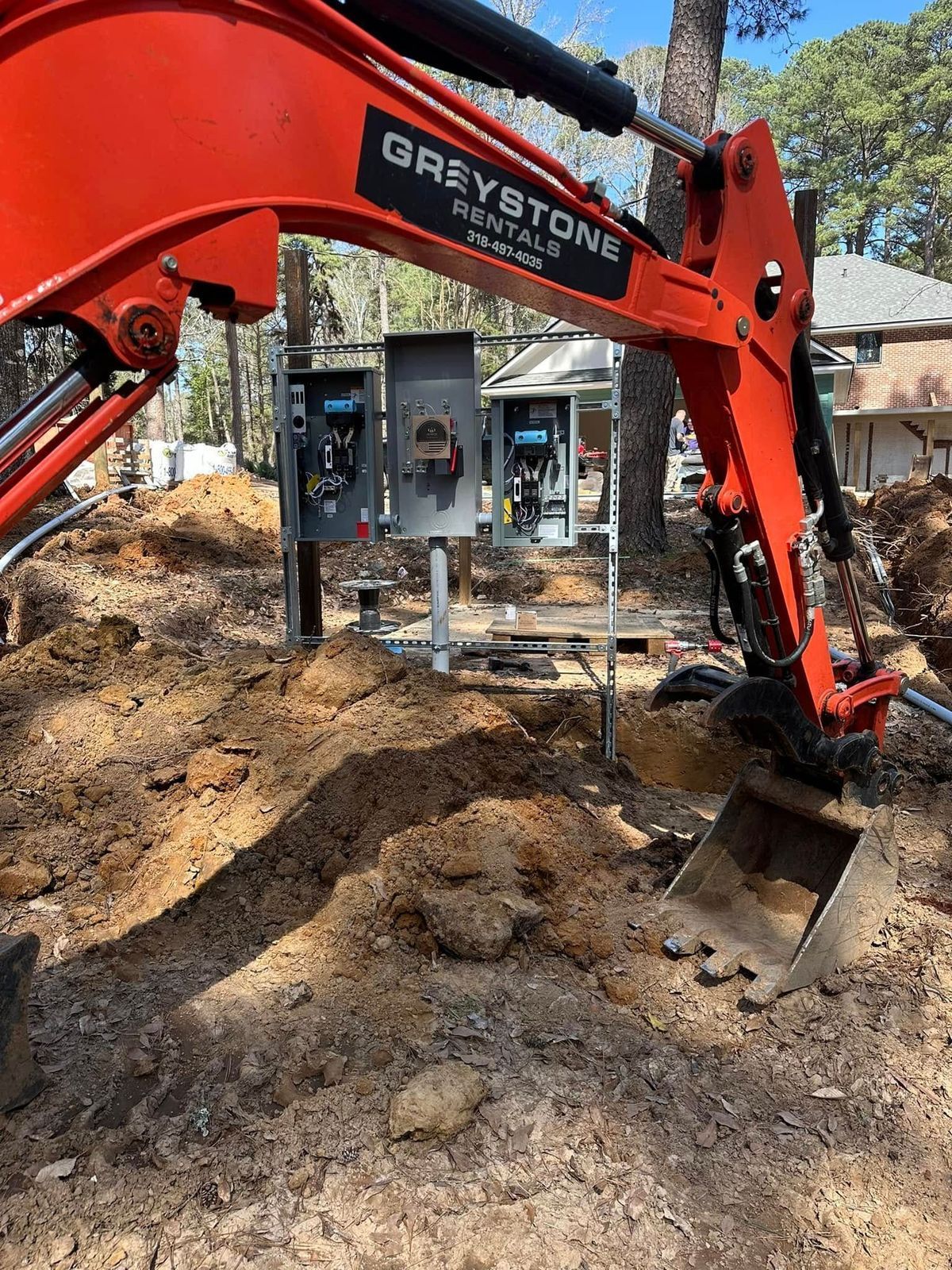 An orange Greystone Rentals excavator digging near electrical boxes in a dirt yard.