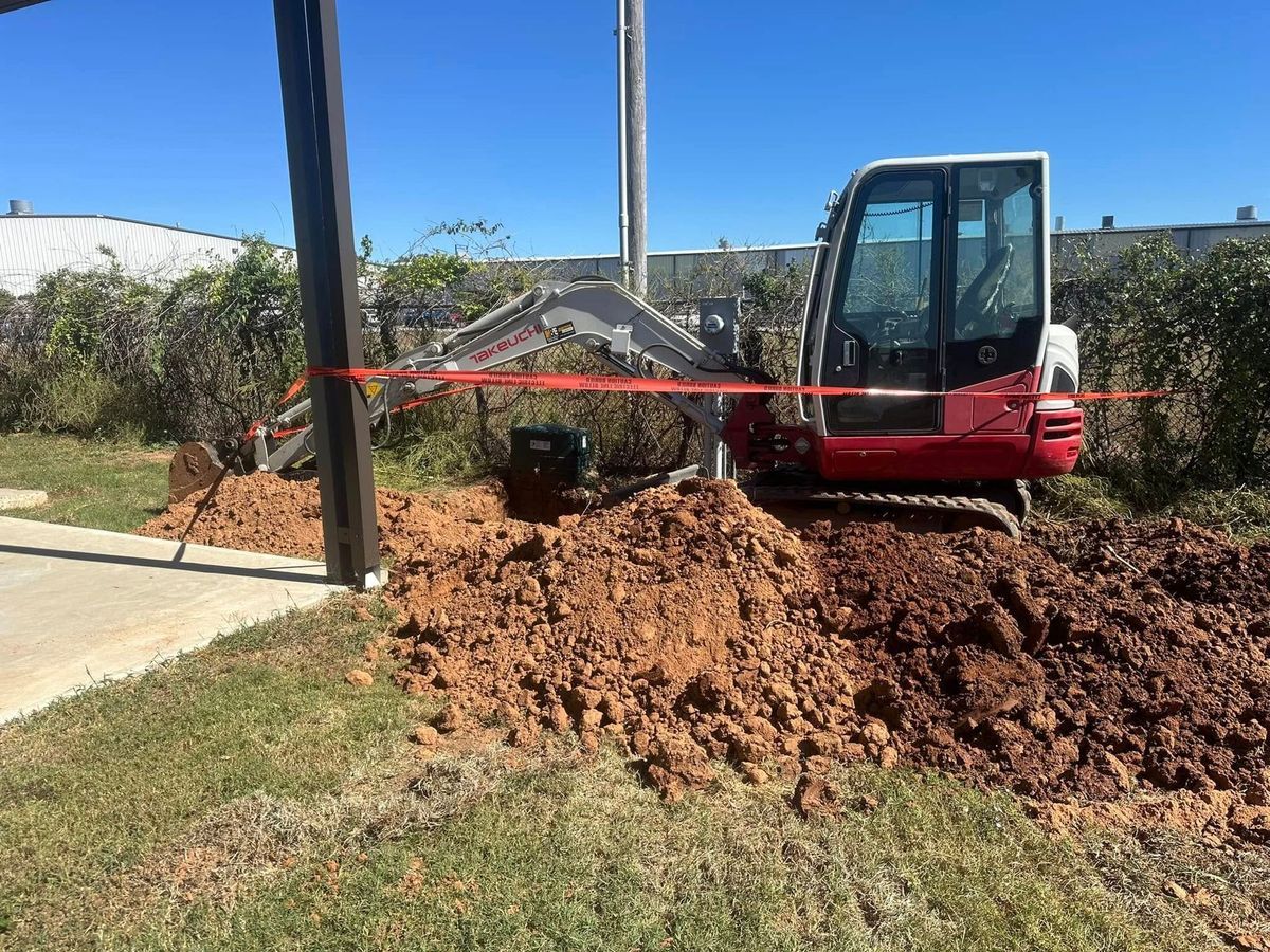 Mini excavator digging near a dark pole and a bush, dirt piled on grass.
