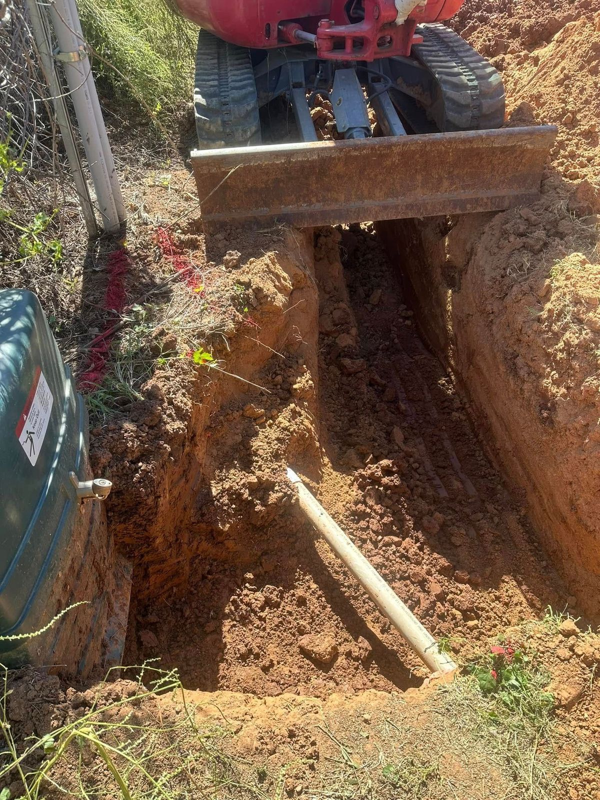 Mini excavator digging a trench in dirt, next to a green tank and fence.