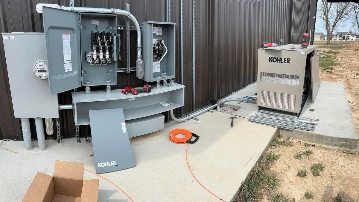 Electrical panel and generator installation next to a metal building, on a concrete pad.