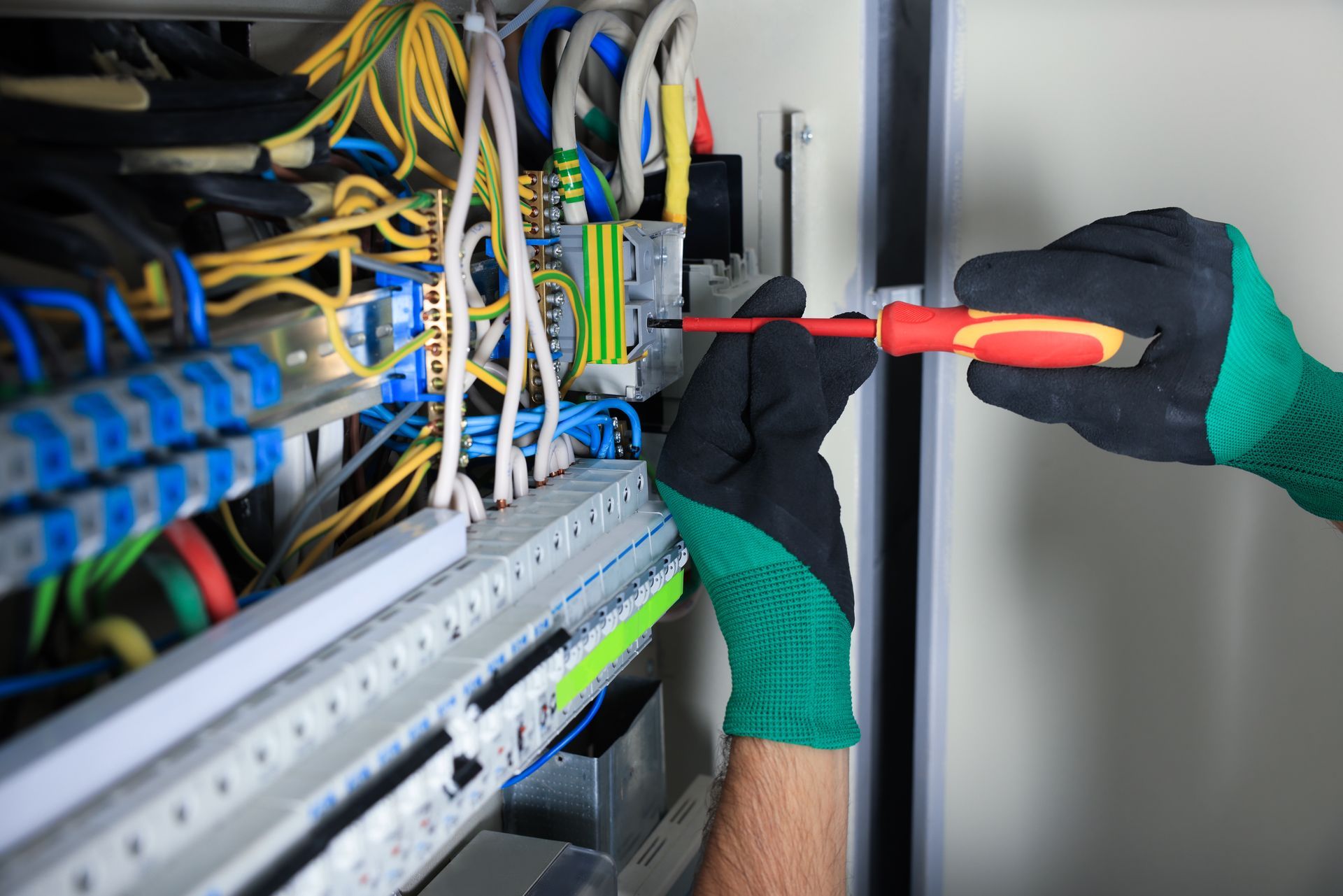 Electrician in gloves uses a screwdriver on electrical panel wiring.