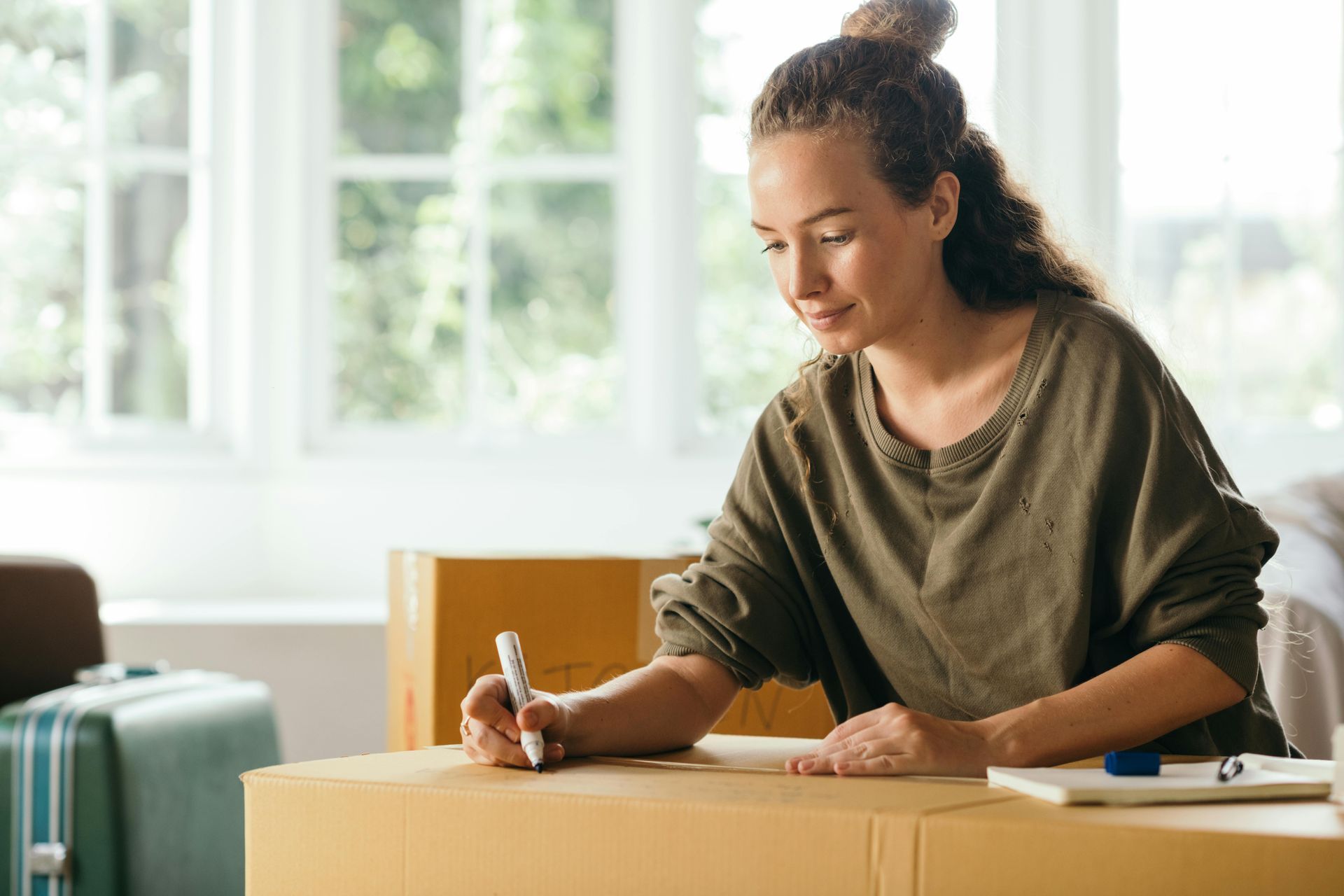 A woman is sitting at a table with moving boxes and writing on a piece of paper.
