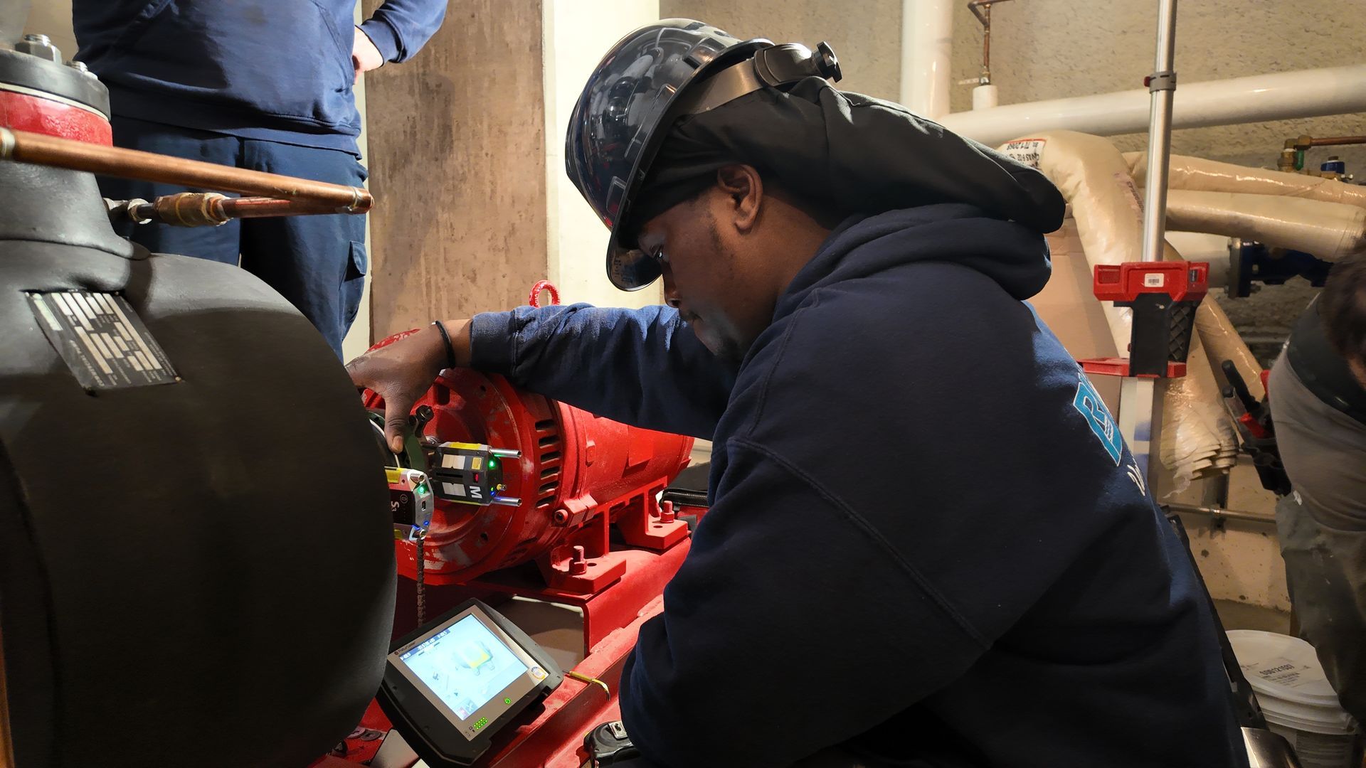 Man in protective gear inspecting red machinery in a mechanical room, using a handheld device.