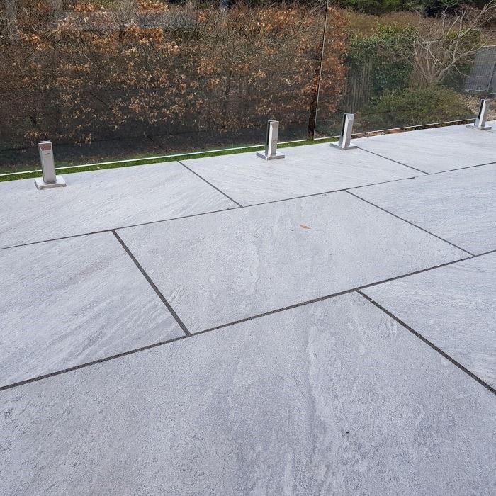 Gray stone patio with metal supports, glass fence, and shrubbery in the background.