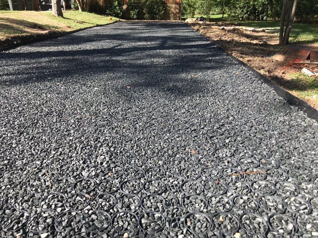 Gravel driveway with black permeable pavers, bordered by dirt and grass, in an outdoor setting.