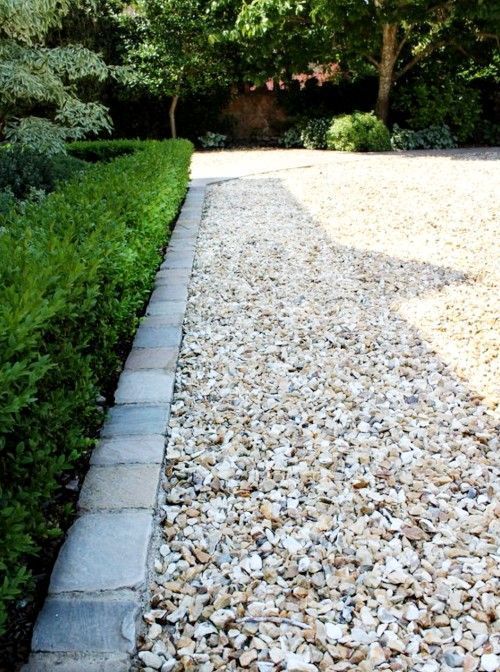 A gravel driveway bordered by a boxwood hedge and stone pavers, leading towards trees.