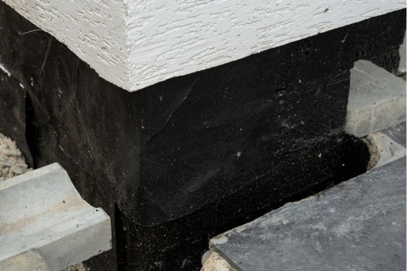 Black waterproof membrane covering the foundation of a building. Gravel and concrete blocks are visible.