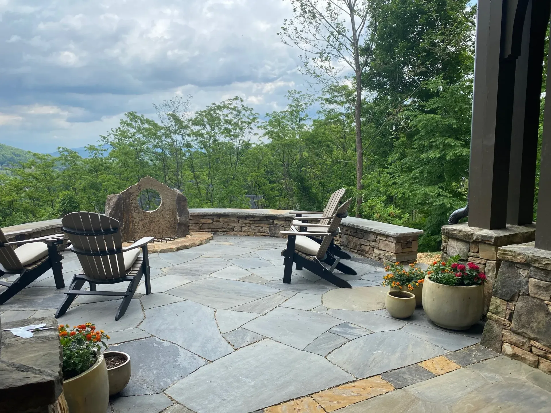 Patio with adirondack chairs, potted flowers, stone wall, and a view of trees and sky.