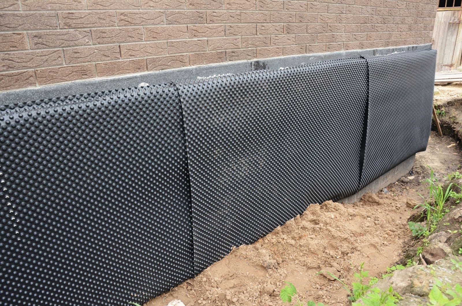 Black dimpled foundation wrap installed against a brick wall, with a sandy soil trench.