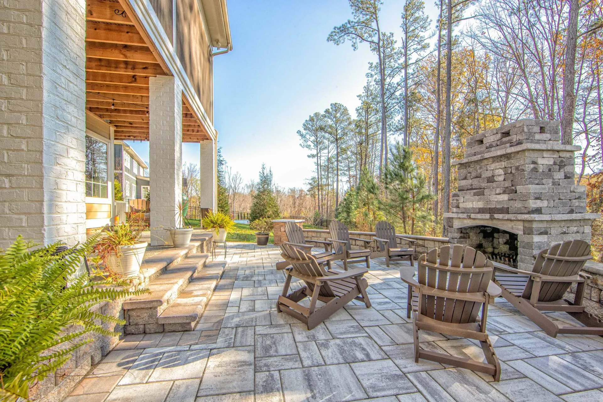 Patio with stone fireplace, wooden chairs, and porch, surrounded by trees on a sunny day.