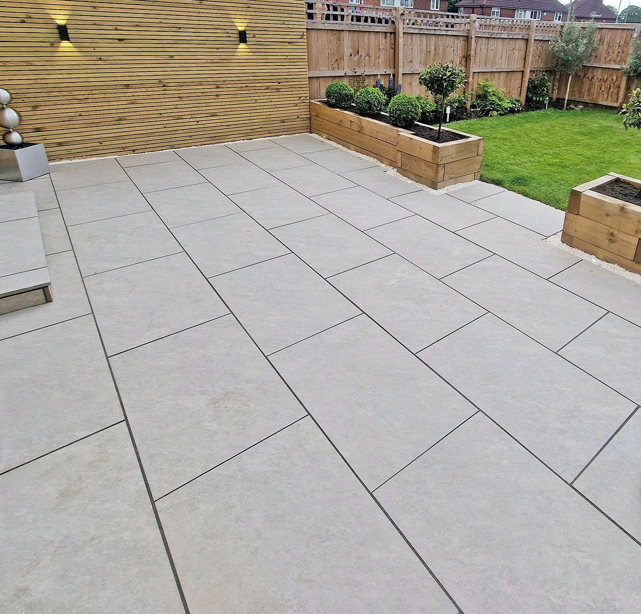 Beige stone patio with rectangular pavers, bordered by a lawn and wooden planters.