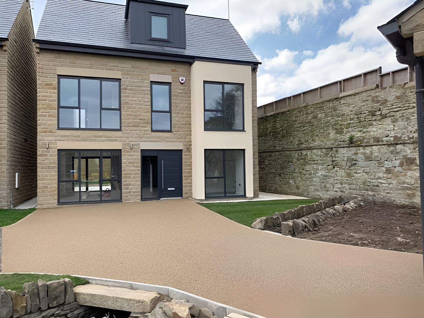 Modern two-story house with stone facade and gravel driveway, next to a stone wall.