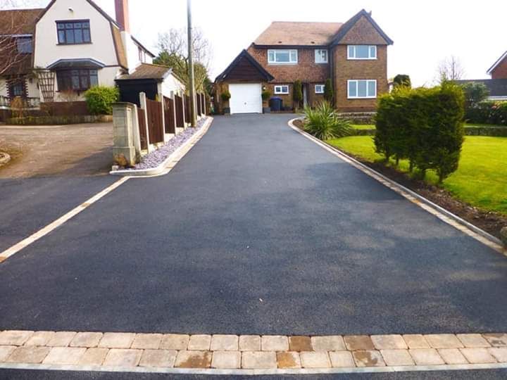 Asphalt driveway leading to a two-story house. Tan borders and a small green bush.