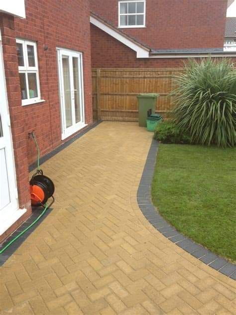 Brick pathway next to a red brick house, leading to a lawn. The pathway has a black border and a green bin is visible.