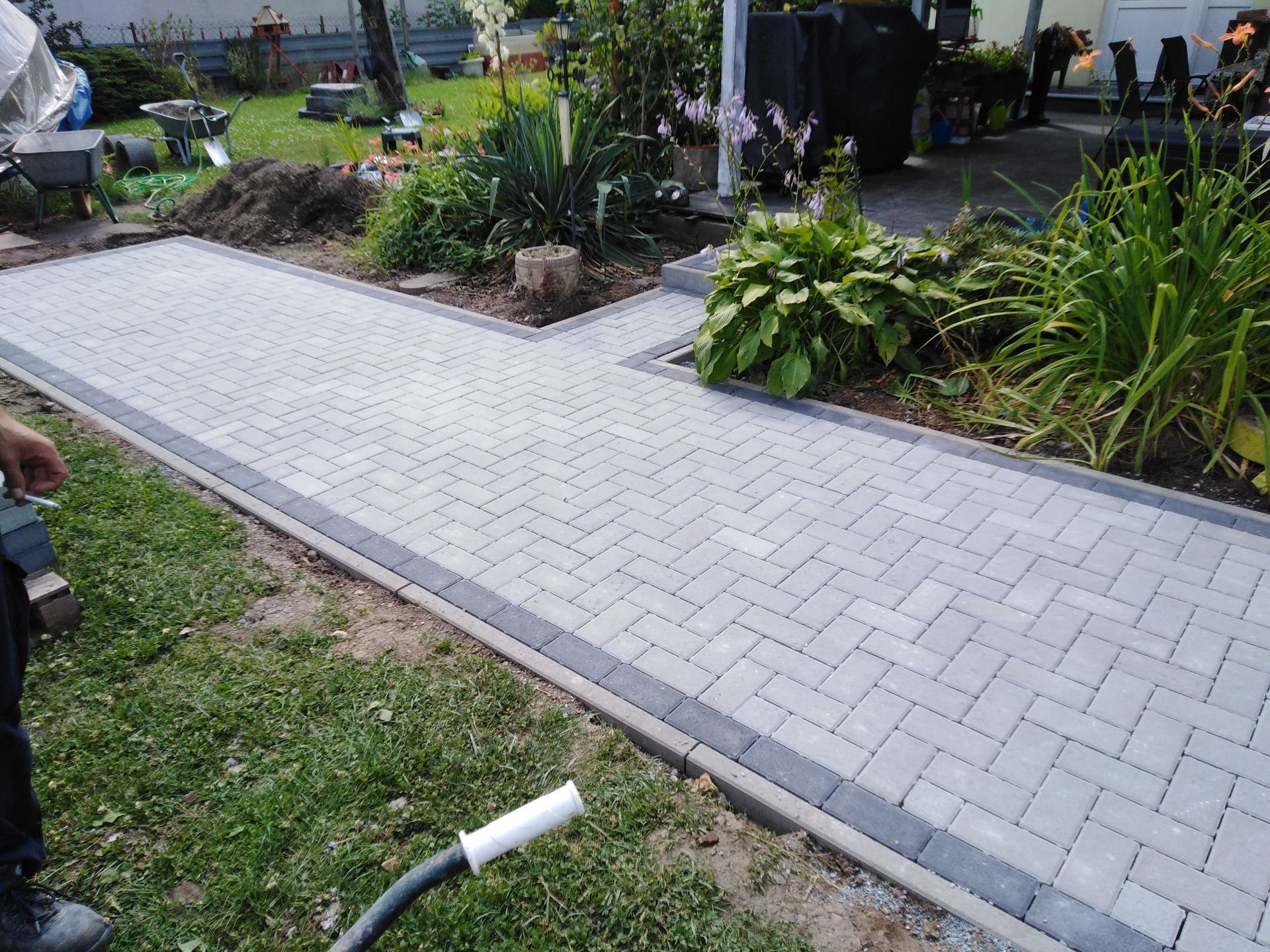 Paved walkway with gray bricks bordered by darker bricks, passing garden with various green plants.