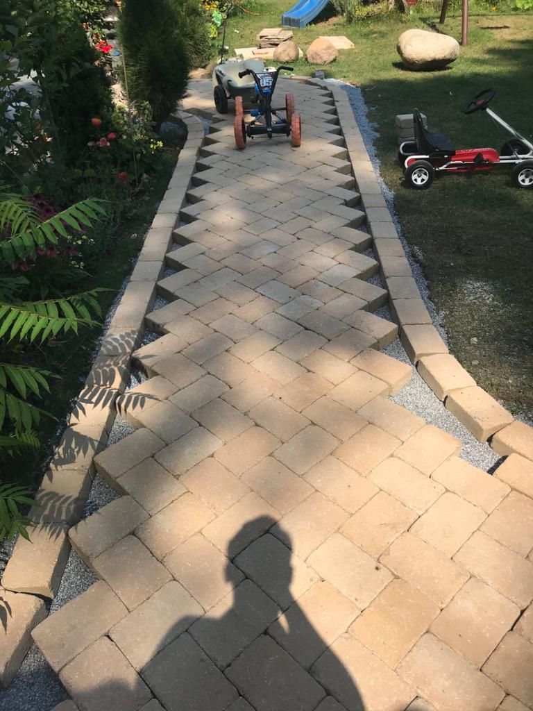 A brick walkway with a herringbone pattern in a garden, bordered by stones.