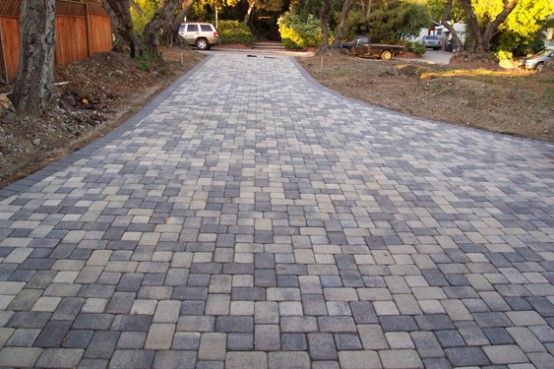 Brick driveway with a mix of gray and tan square pavers, leading towards trees and a vehicle.