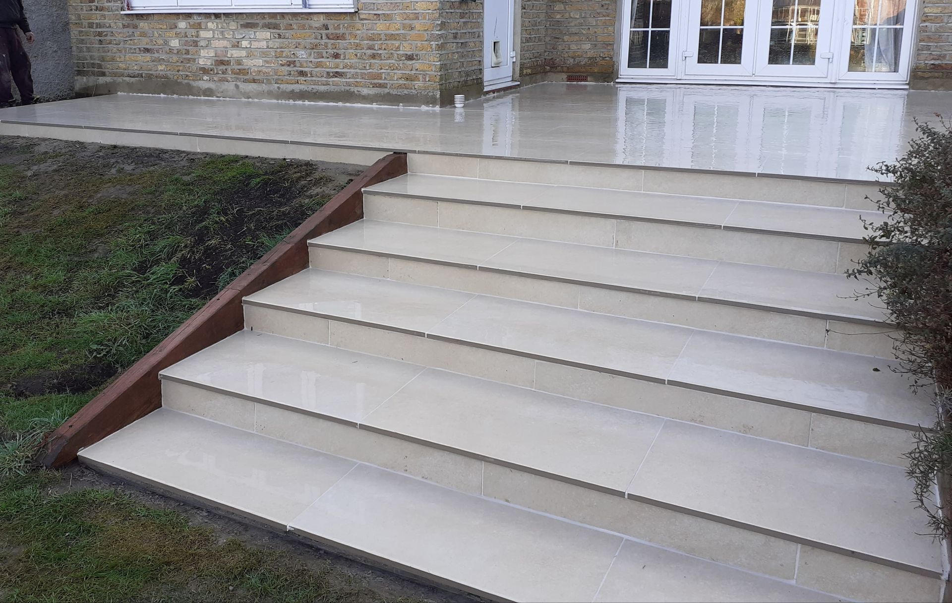 Stone steps lead up to a patio and a building with glass doors; brown retaining wall on one side.