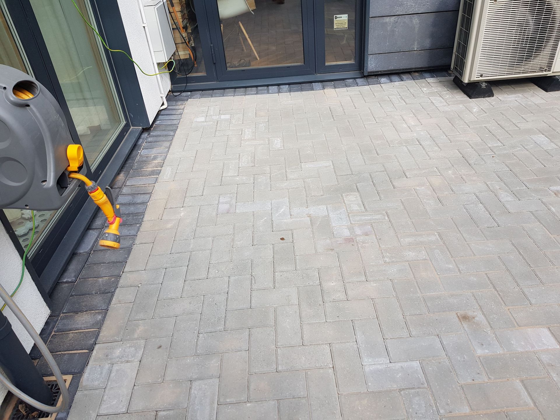 Gray brick patio with a hose reel, next to a building with glass doors and an air conditioning unit.