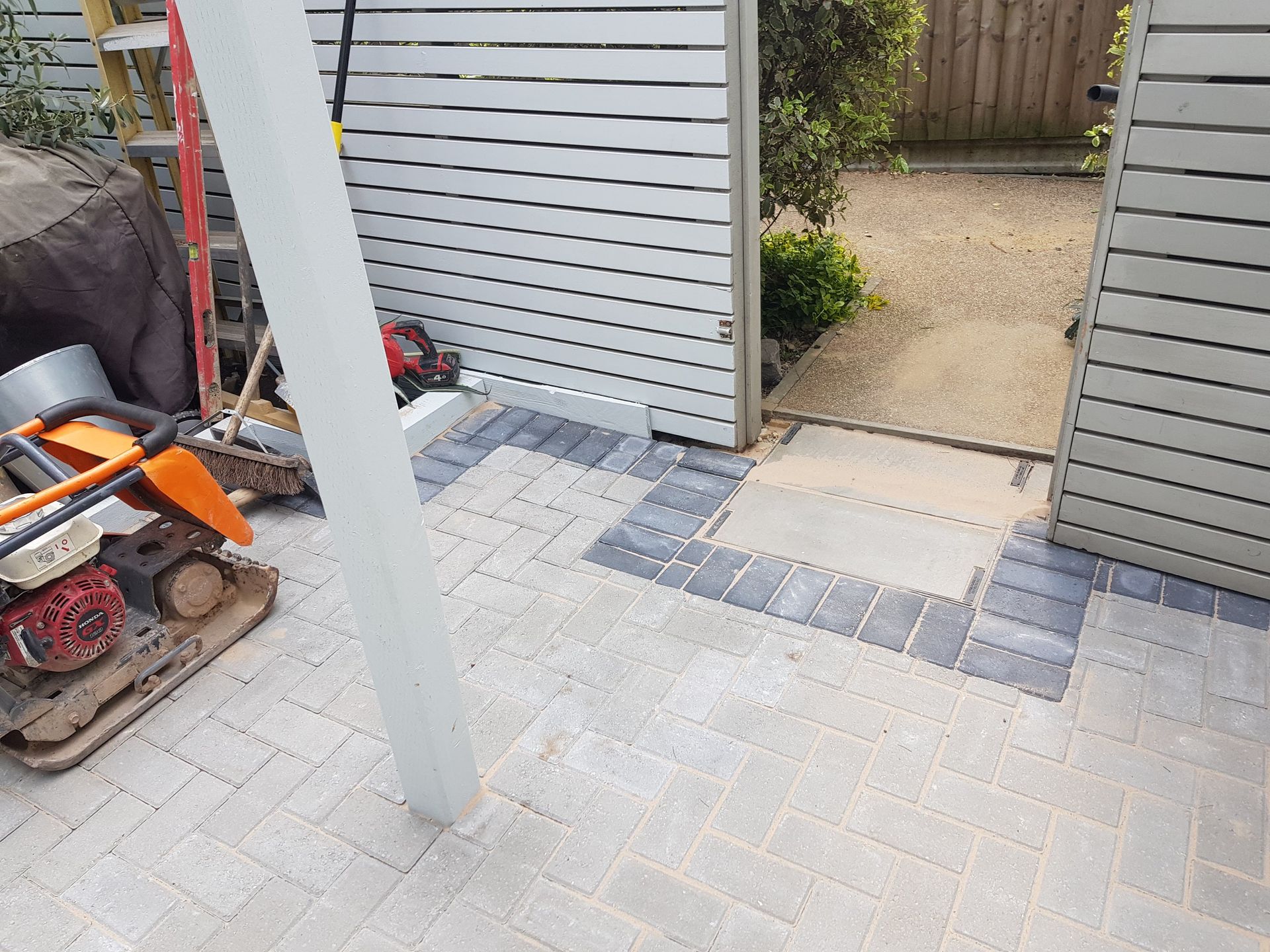 Newly paved patio with grey and beige bricks, leading to a gravel pathway, tools on left.