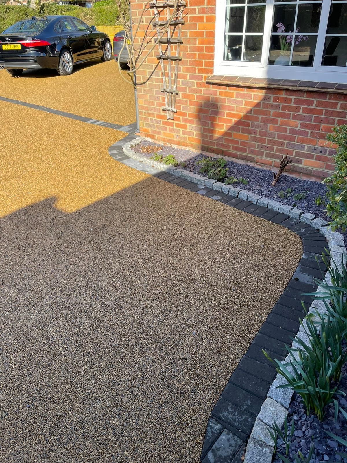 Driveway with dark gravel and gold gravel, bordered with brick and stone. Brick house with a shadow.