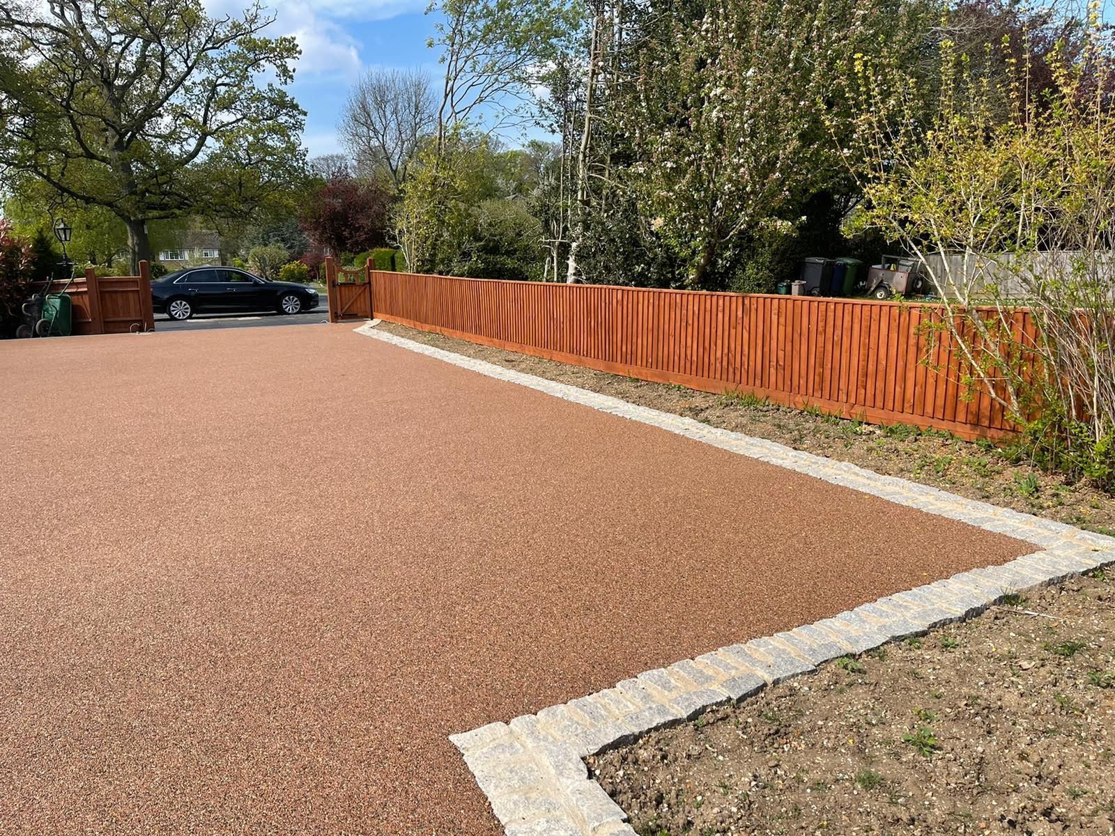 Brown gravel driveway with a wooden fence, concrete edging, and a black car in the background.
