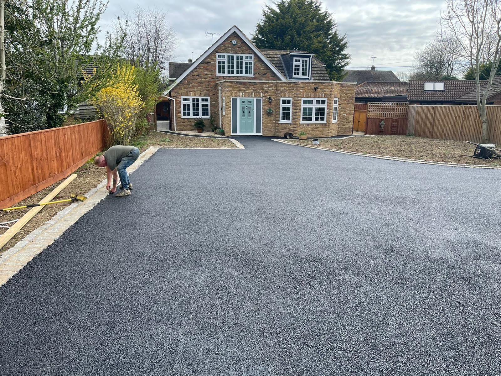 Man working on a dark driveway in front of a light-colored brick house with a blue door; cloudy day.