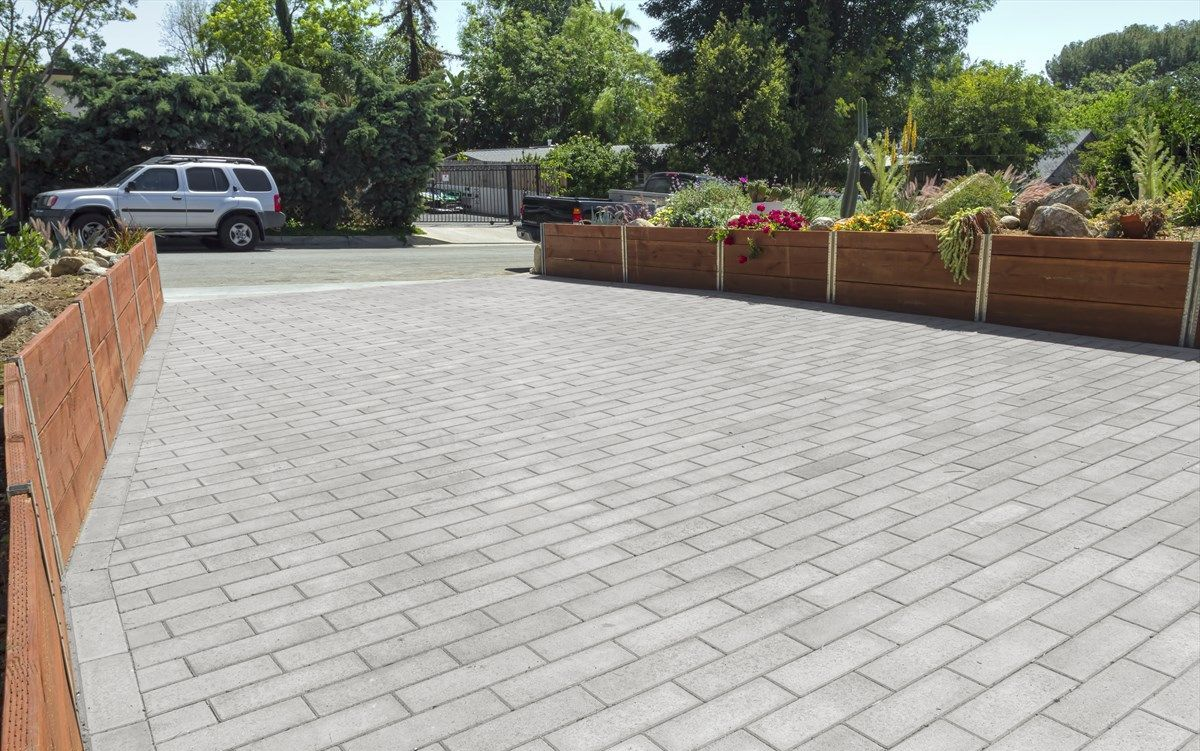 Brick paved driveway with a planted wooden border; a parked SUV and trees in the background.