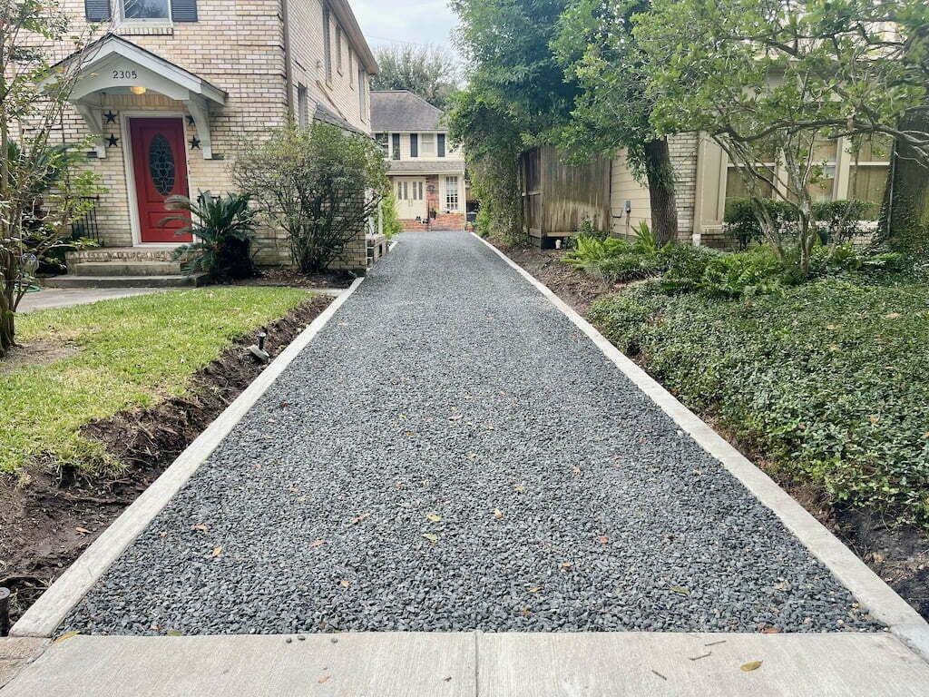 Two-story brick house with white gravel driveway, gray garage doors, and a blue sky.