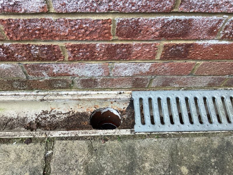 Brick wall with a drainage channel containing debris, next to a metal grate.