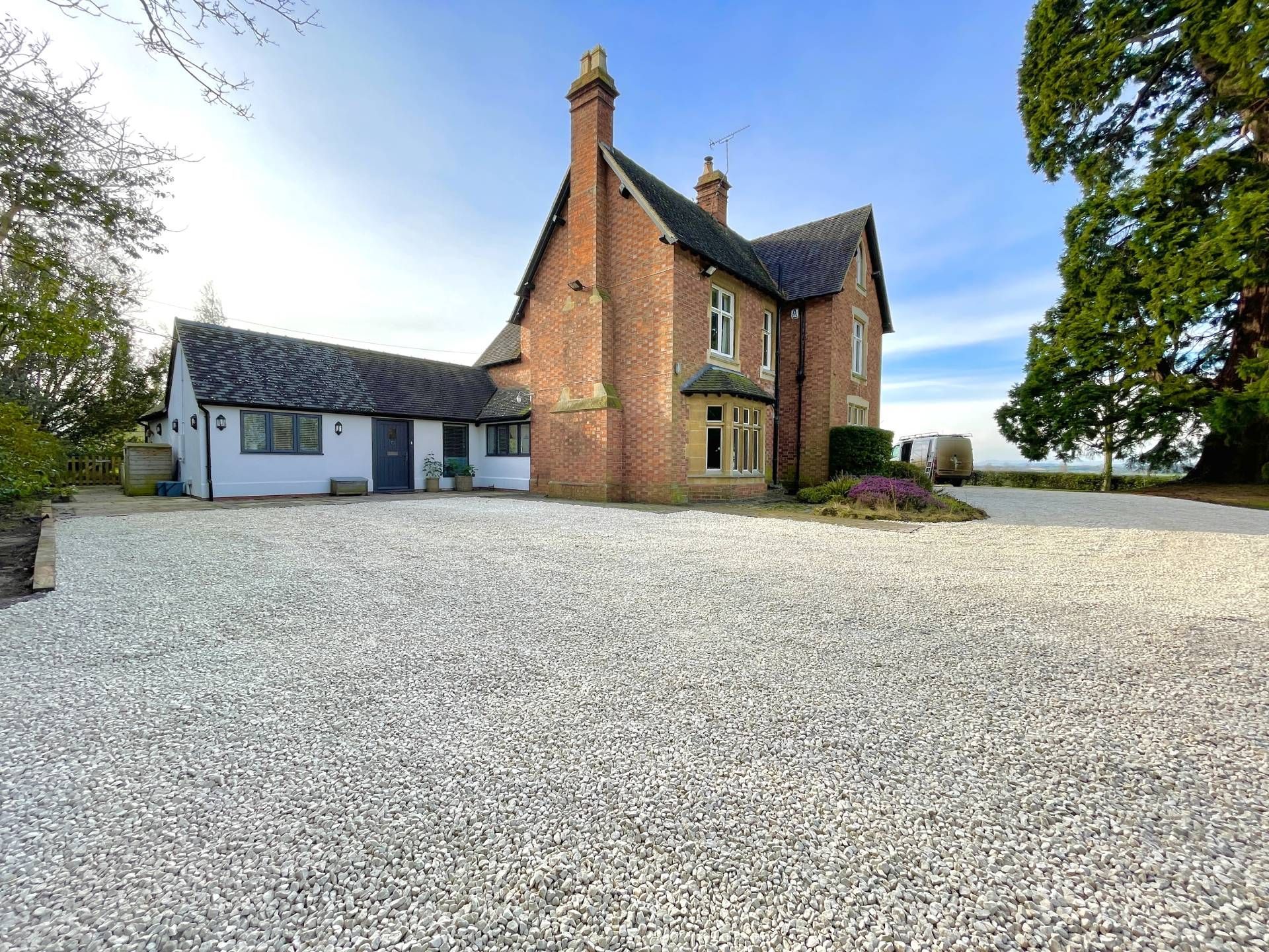 A brick house with white gravel driveway, a blue door, and a large tree on a sunny day.