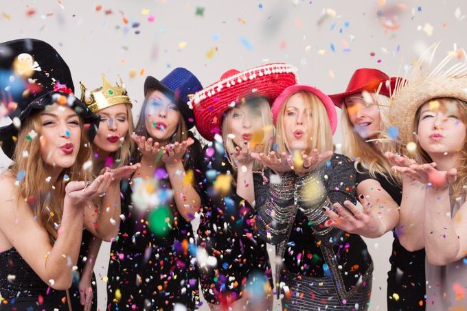 A group of people in colorful party hats blow confetti toward the camera against a white background.