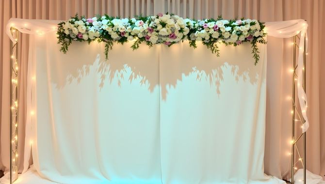 A wedding backdrop featuring a white draped curtain with a laser-cut pattern, adorned with a floral garland and fairy lights.