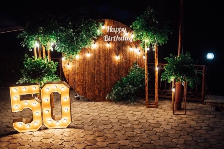 A 50th birthday party display with large, illuminated number 50 lights, a circular wooden backdrop, and greenery at night.