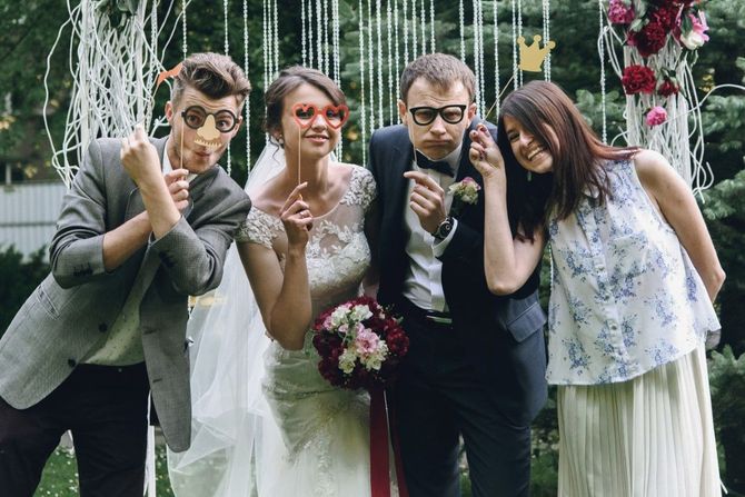 Four people pose outdoors with whimsical photo props in front of a white, vine-decorated backdrop.