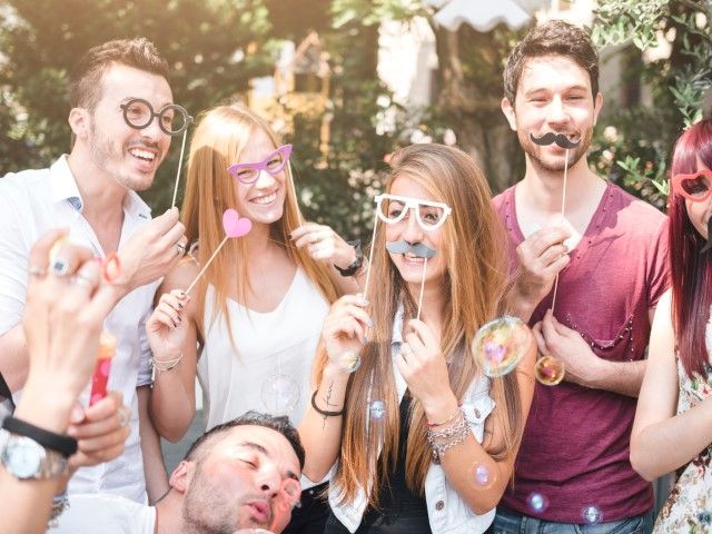 A group of friends posing outdoors with fun props like fake mustaches and colorful glasses at a casual party.