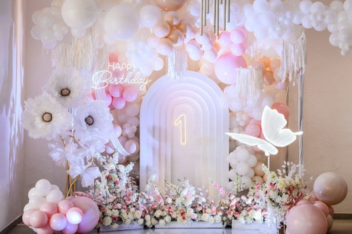 A first birthday party backdrop featuring an arched white structure with a glowing number one, balloons, and floral decor.