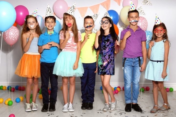 Seven children wearing party hats stand in a row against a white wall decorated with colorful balloons and streamers.
