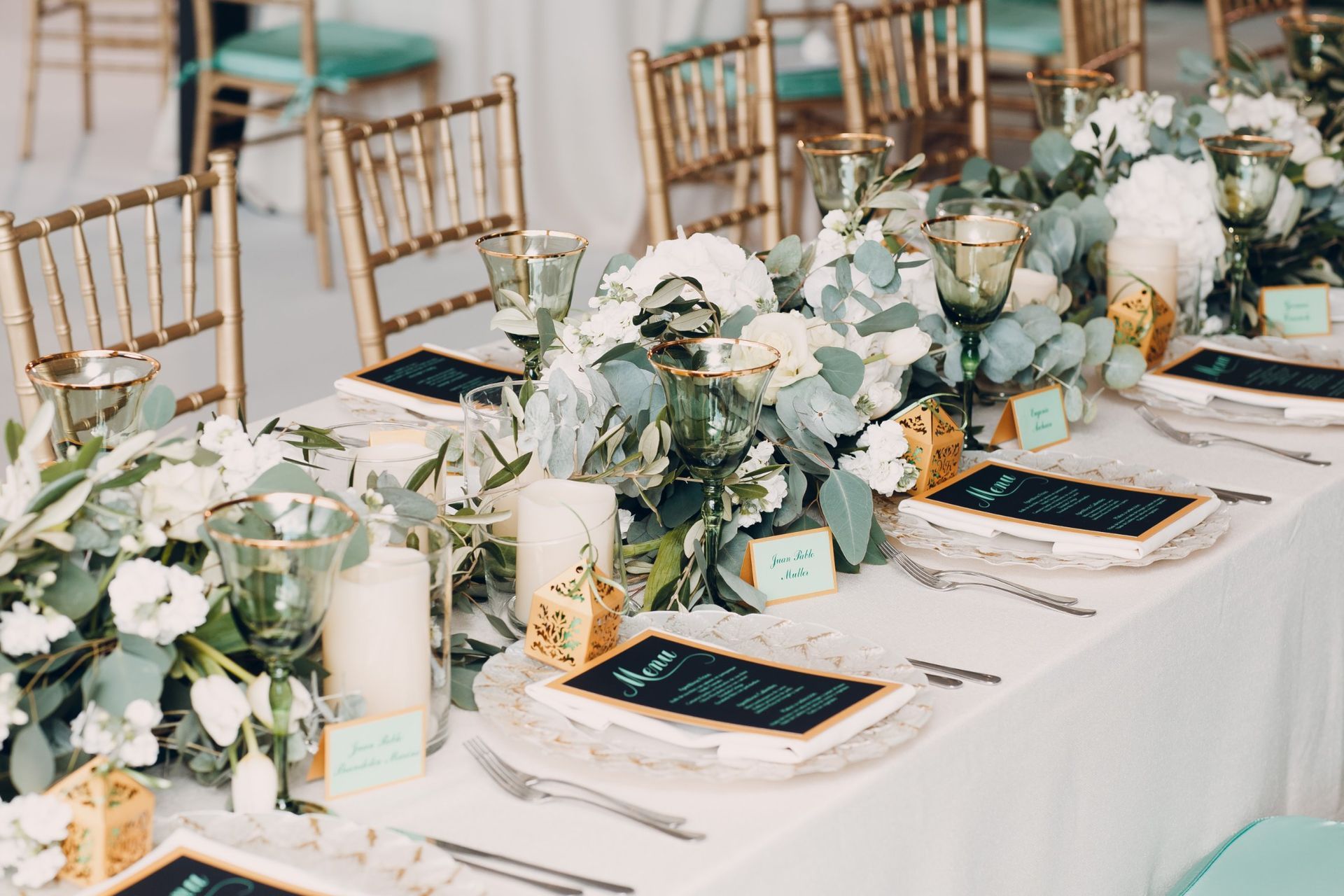 An elegant wedding table setting with gold chairs, white floral centerpieces, greenery, candles, and black menu cards.