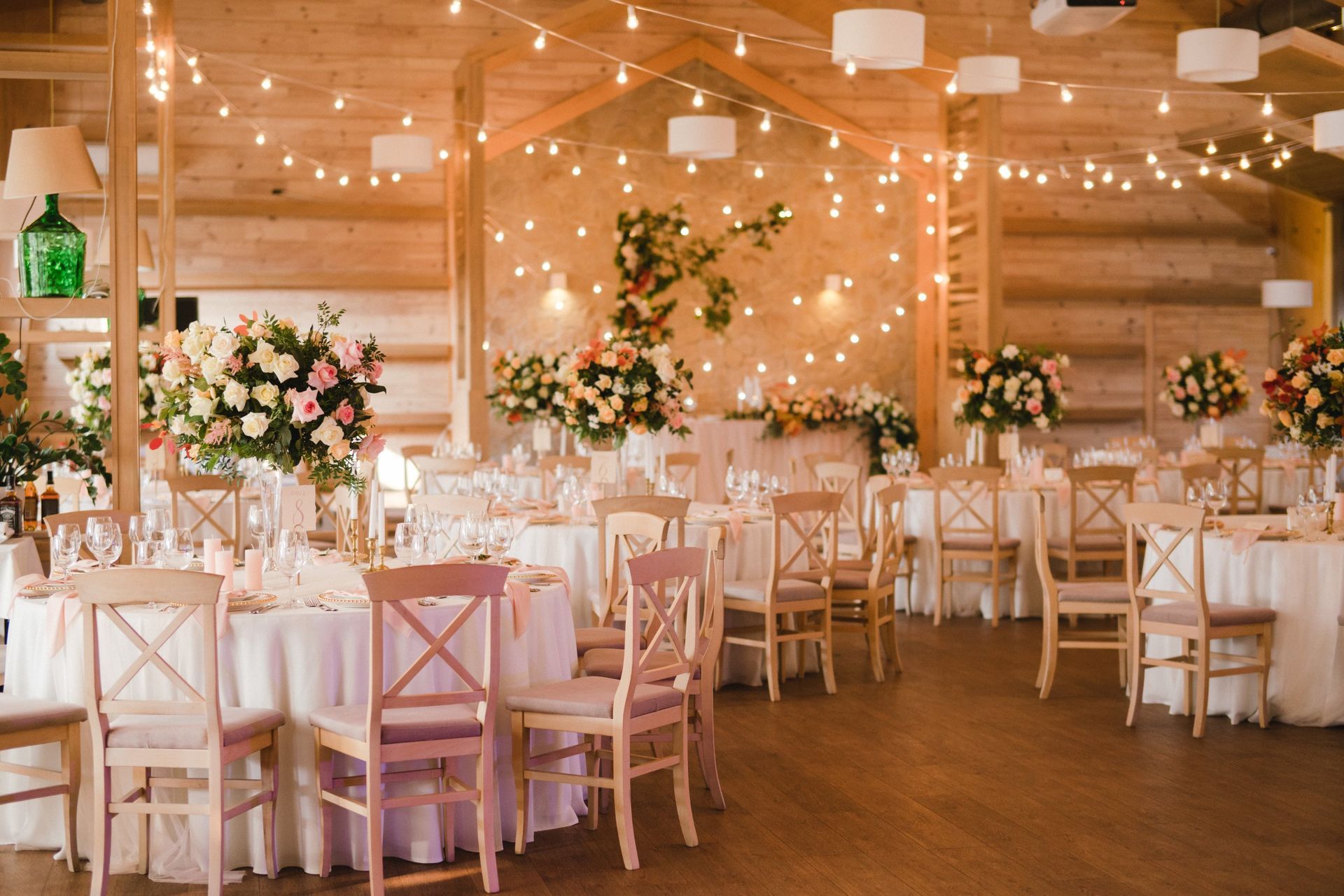 Round dining tables set for a wedding reception in a rustic wooden room decorated with warm string lights and flowers.