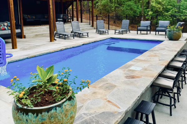 Rectangular blue swimming pool with stone deck, patio chairs, and potted plants in a courtyard