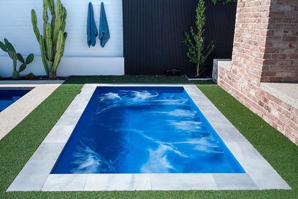 Small rectangular backyard pool with blue water, white border, green turf, and brick wall backdrop