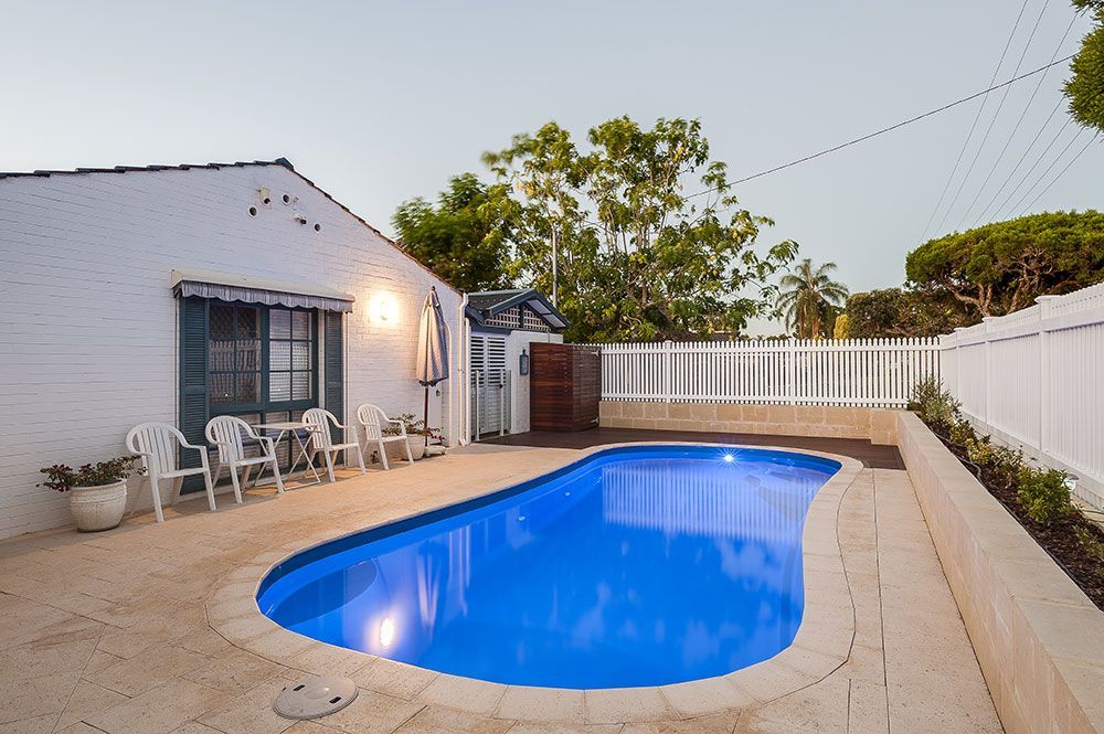 Backyard with a lit blue swimming pool, patio chairs, and a white fence at dusk